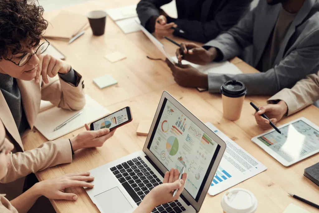 A team meeting with laptops and tablets displaying data visuals. Coffee cups and notepads scattered across a wooden table.