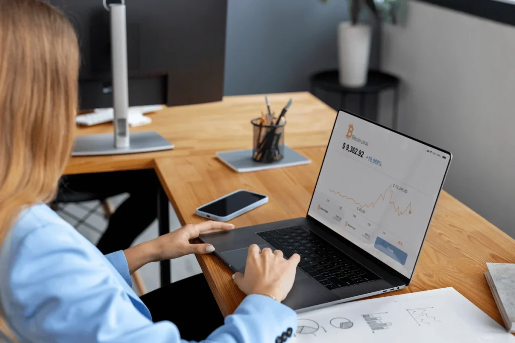 A person in a blue blazer analyzes Bitcoin price data on a laptop in a modern office setting with a smartphone nearby.