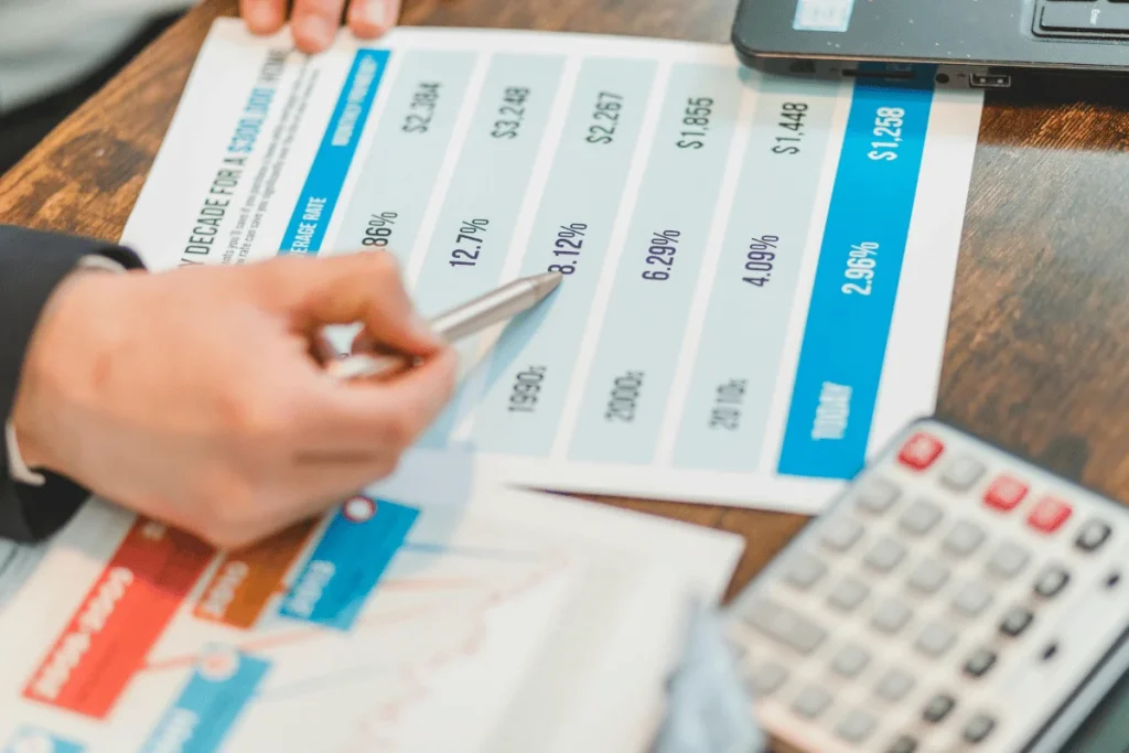 A person's hand holding a pen points to a financial chart on paper, displaying historical data and interest rates, alongside a calculator and laptop.