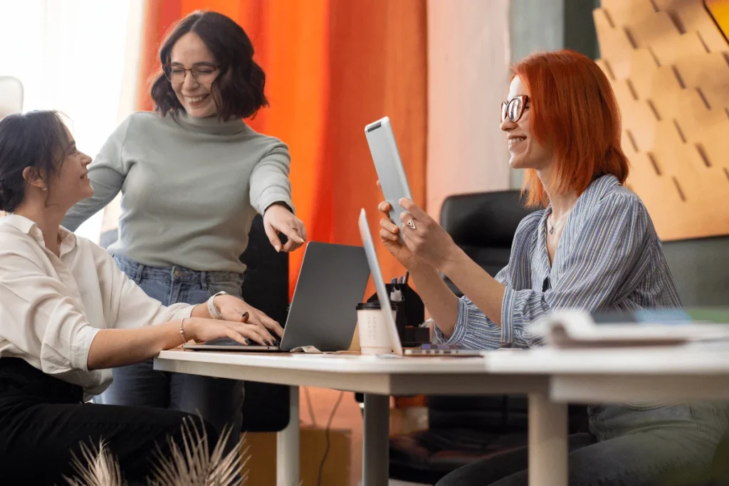 Three women in an office collaborate cheerfully. One stands pointing at a laptop, while others sit, one typing, the other holding a tablet, smiling.