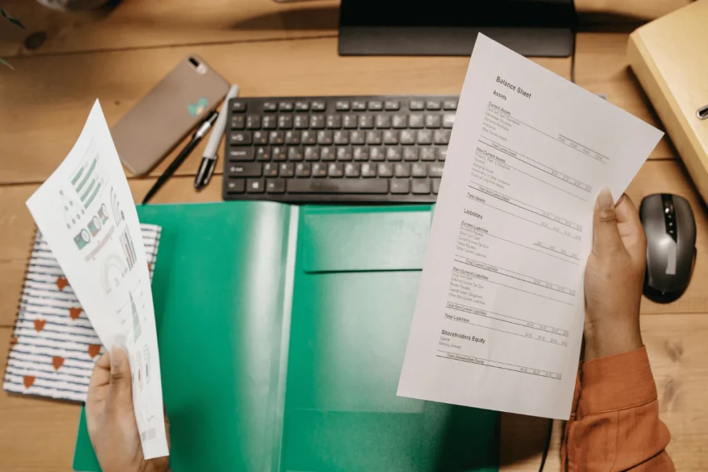 A person holds two financial documents over a desk with a green folder, keyboard, and computer mouse in the background.