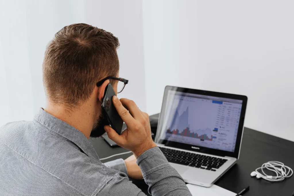 A man with glasses is on a phone call, focusing on financial charts displayed on his laptop screen.