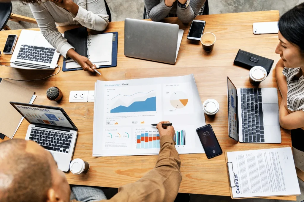 A collaborative meeting around a wooden table, displaying laptops, coffee cups, and business charts for analysis and discussion.