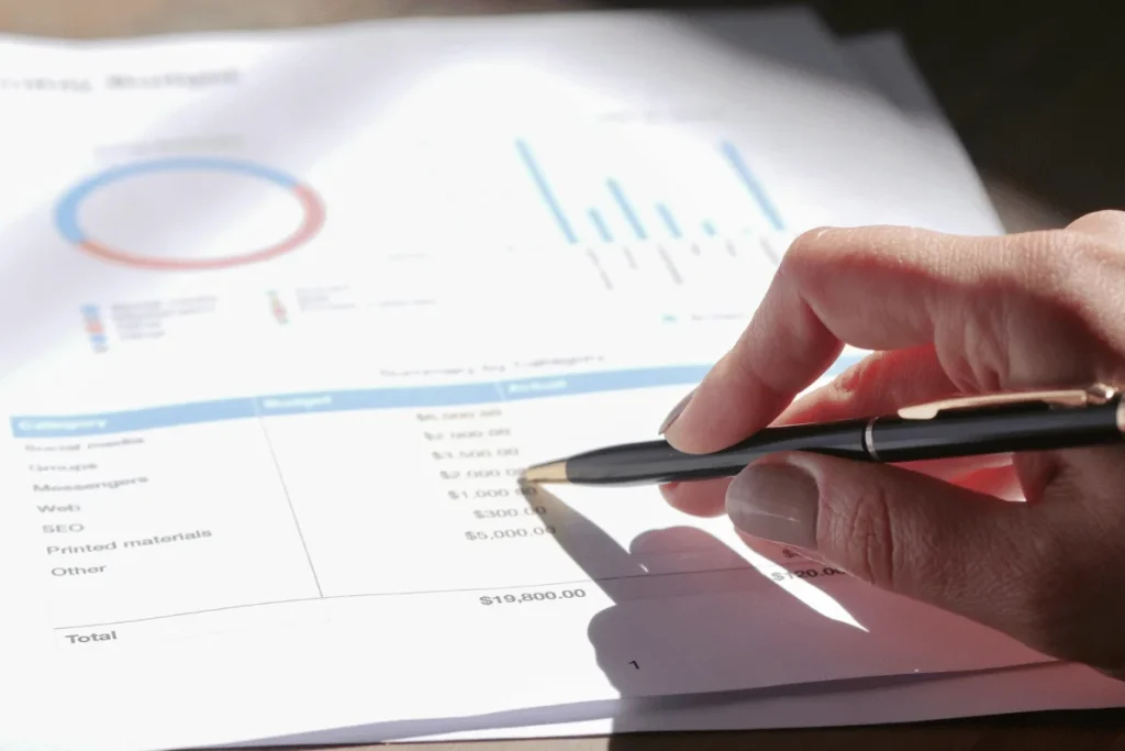 A hand holds a pen over a printed budget document with graphs and financial categories, indicating a focus on budgeting analysis.