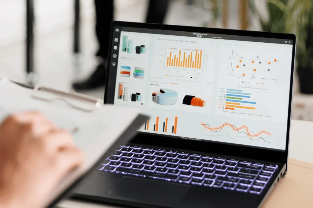 Laptop displaying various charts and graphs, including bar, pie, and line charts, signifies data analysis. Clipboard held by a person in the foreground.