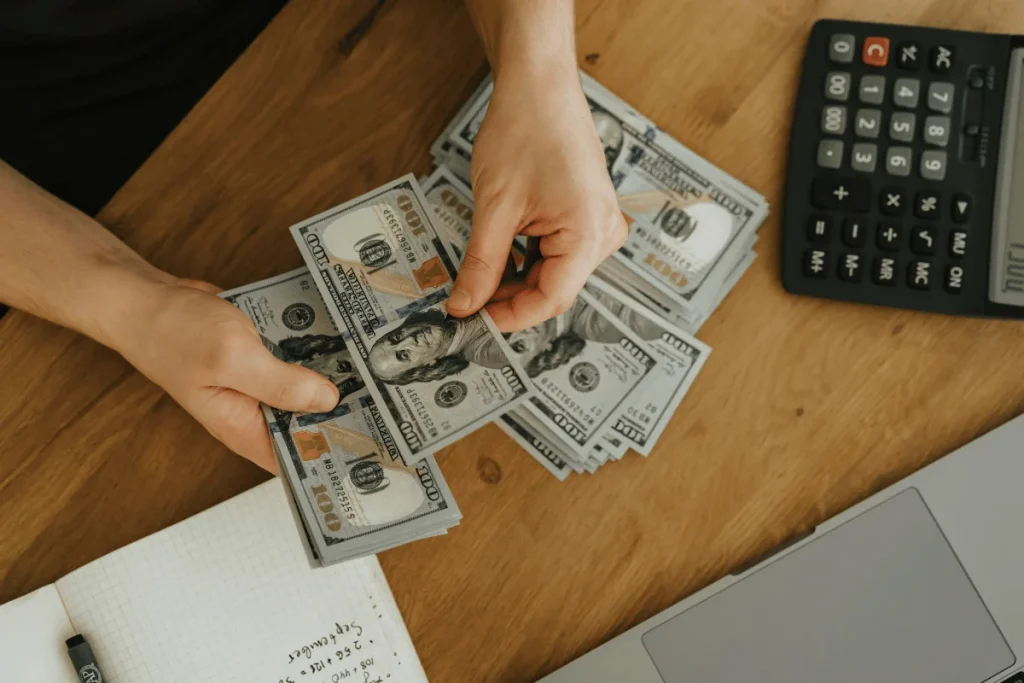 Hands counting numerous $100 bills on a wooden table. Nearby, there's an open notebook, a calculator, and part of a laptop, implying financial planning.