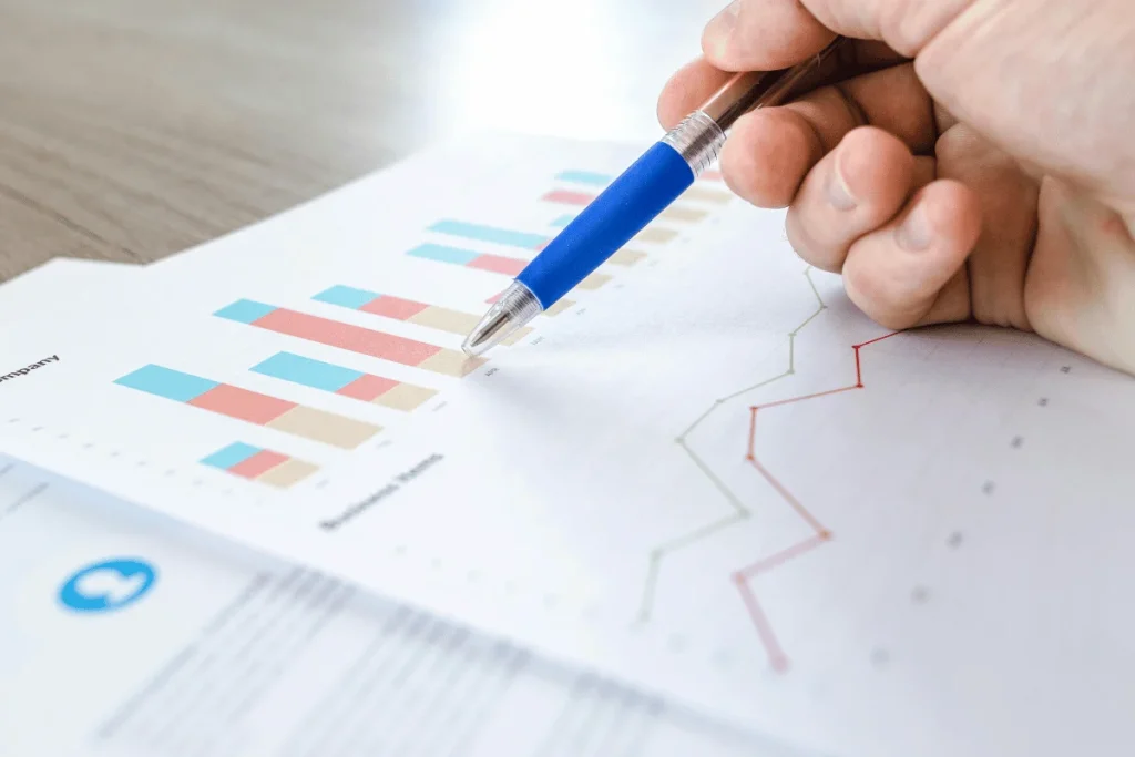 A hand holds a blue pen, pointing at colorful bar and line graphs on paper. The image suggests data analysis or business strategy review.
