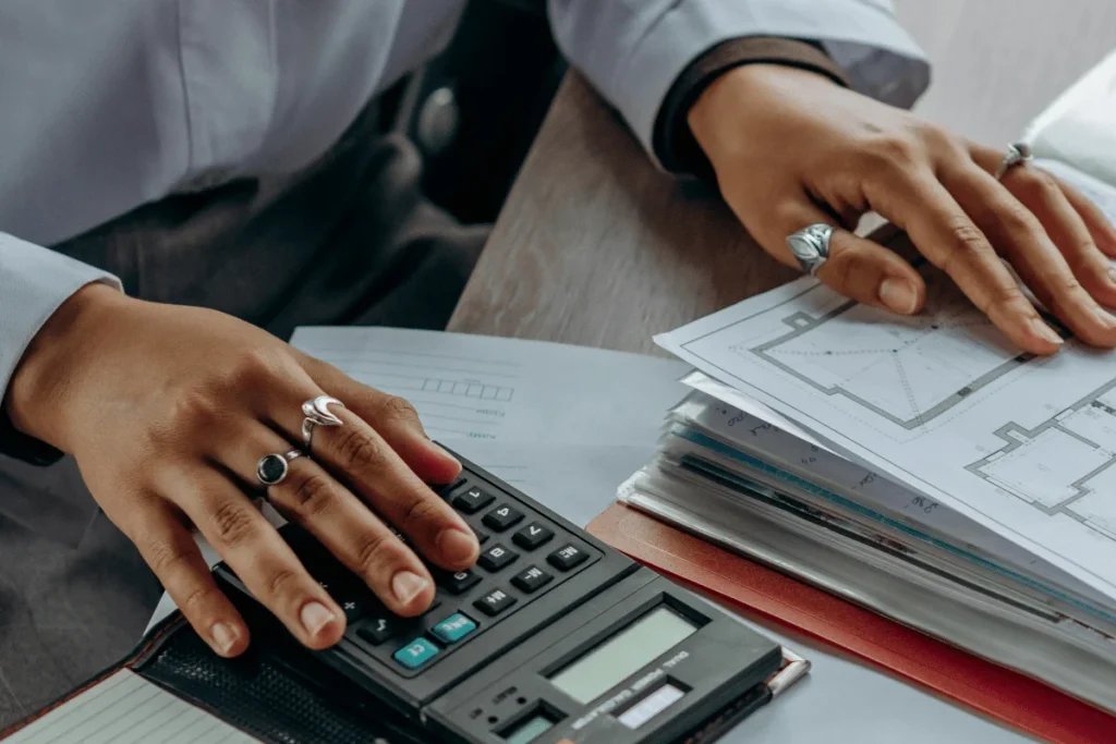 Hands of a person with rings working on architectural blueprints; one hand uses a calculator, conveying focus and precision in design work.