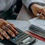 Hands of a person with rings working on architectural blueprints; one hand uses a calculator, conveying focus and precision in design work.