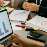 Business meeting scene with hands on a table, featuring charts and graphs on papers and a laptop. The atmosphere is focused and collaborative.