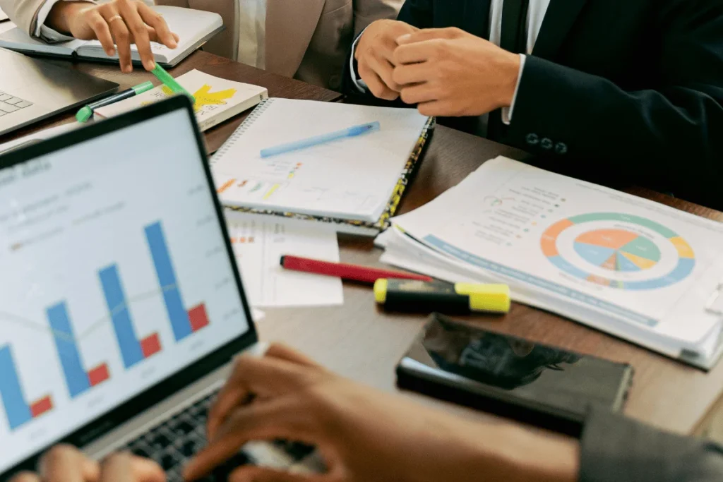People in business attire reviewing charts and graphs on a laptop and papers in a meeting. Pens and a highlighter are scattered on the table.