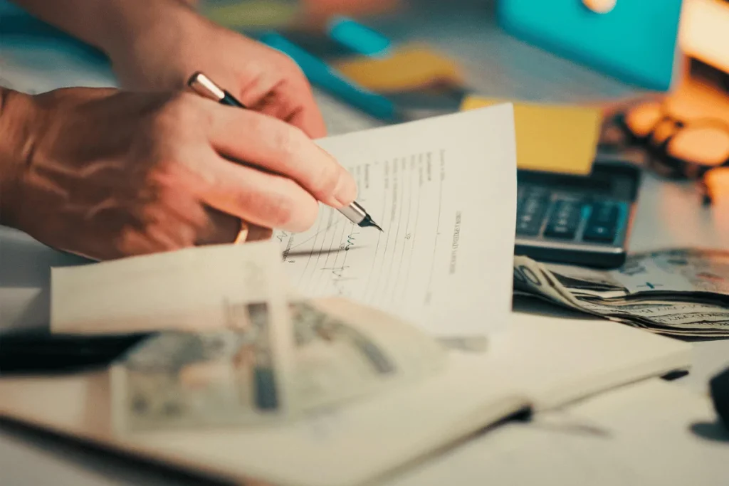 Hand signing financial documents with cash and calculator on the desk.