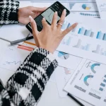 A person in a plaid jacket analyzes data on a smartphone surrounded by charts and graphs.