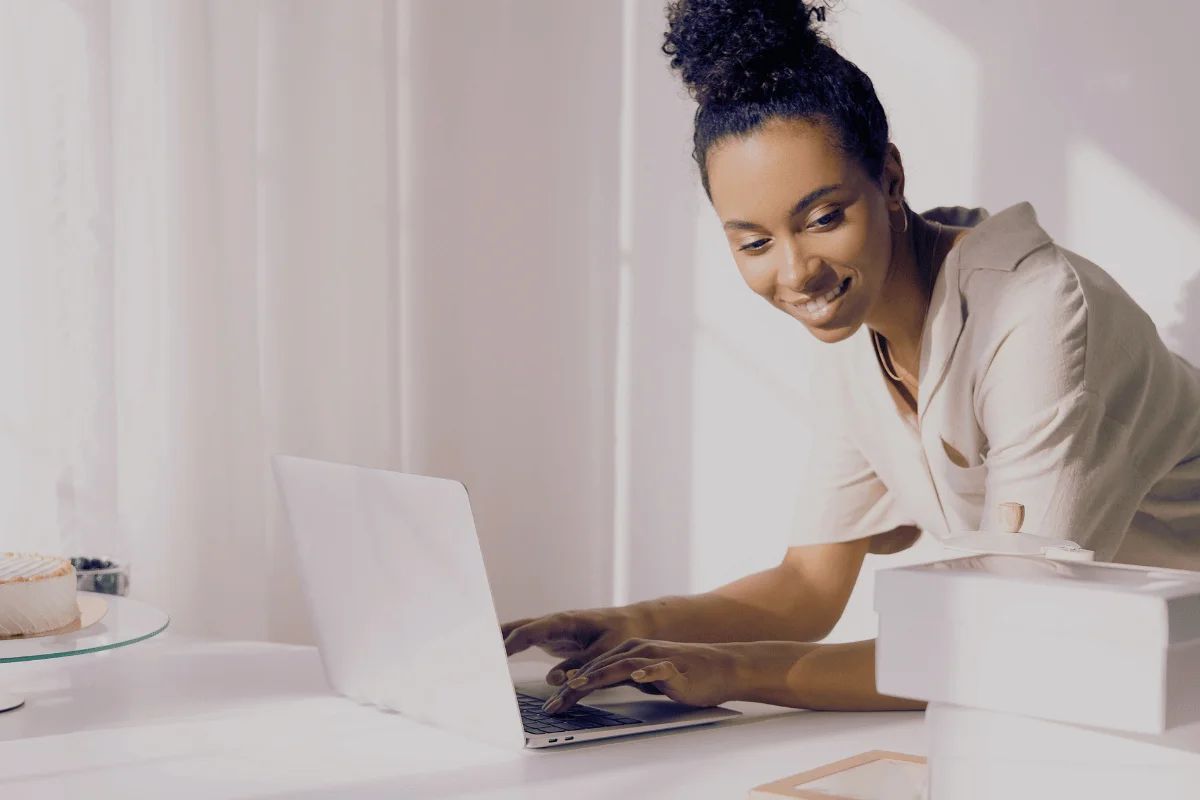A person typing on a laptop at a desk, with a cake on a glass stand and boxes nearby, in a bright, softly lit room.