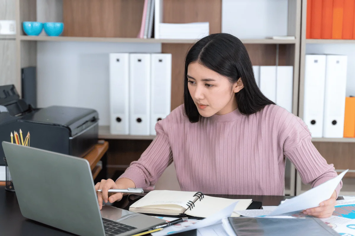 A young woman in a pink sweater focuses on a laptop at a desk, surrounded by papers and an open notebook.
