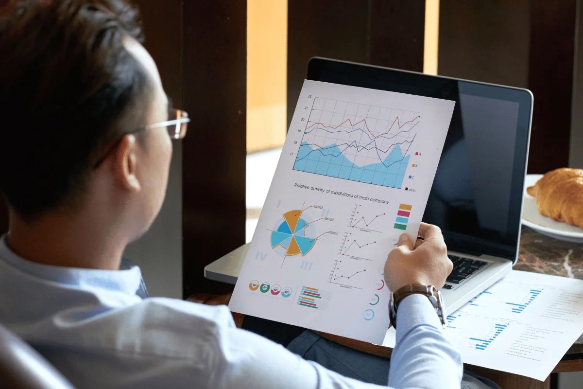 A man in a light blue shirt reviews graphs and charts on a document and laptop, displaying financial data.
