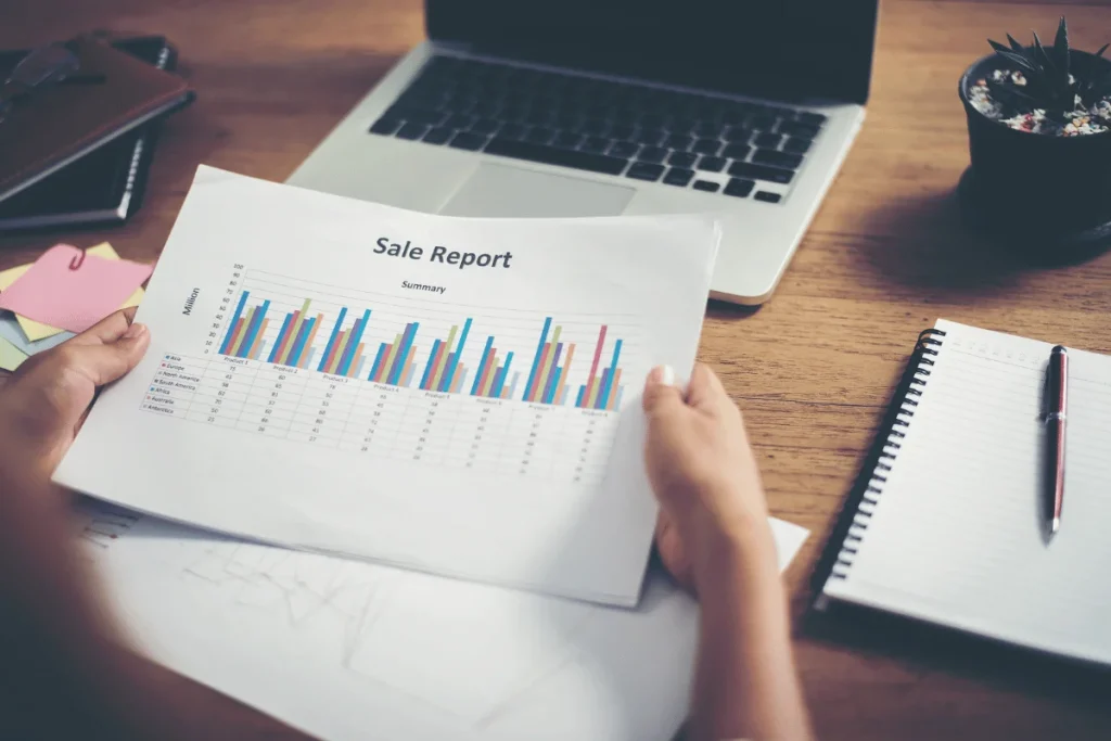 A person holds a sale report with colorful graphs and data, sitting at a desk with a laptop, notepad, and sticky notes.