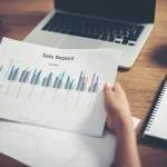 A person holds a sale report with colorful graphs and data, sitting at a desk with a laptop, notepad, and sticky notes.