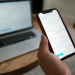 A person holds a smartphone displaying stock market data, with a laptop showing graphs in the background, on a wooden table.