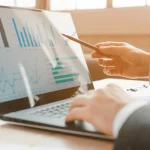 A person in a suit uses a laptop displaying bar and line charts.