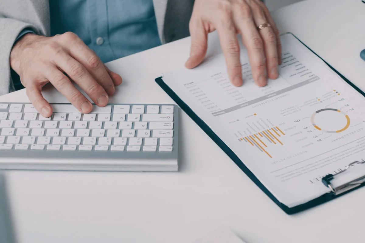 Hands of a person in a suit organize papers with graphs on a clipboard beside a white computer keyboard, depicting focus and organization in an office setting.