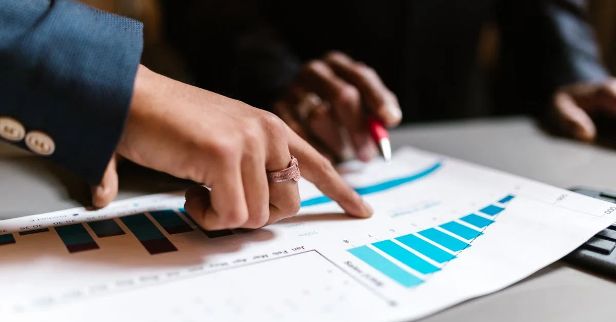Two hands point at a financial graph with blue bars on a printed report, suggesting a business discussion.