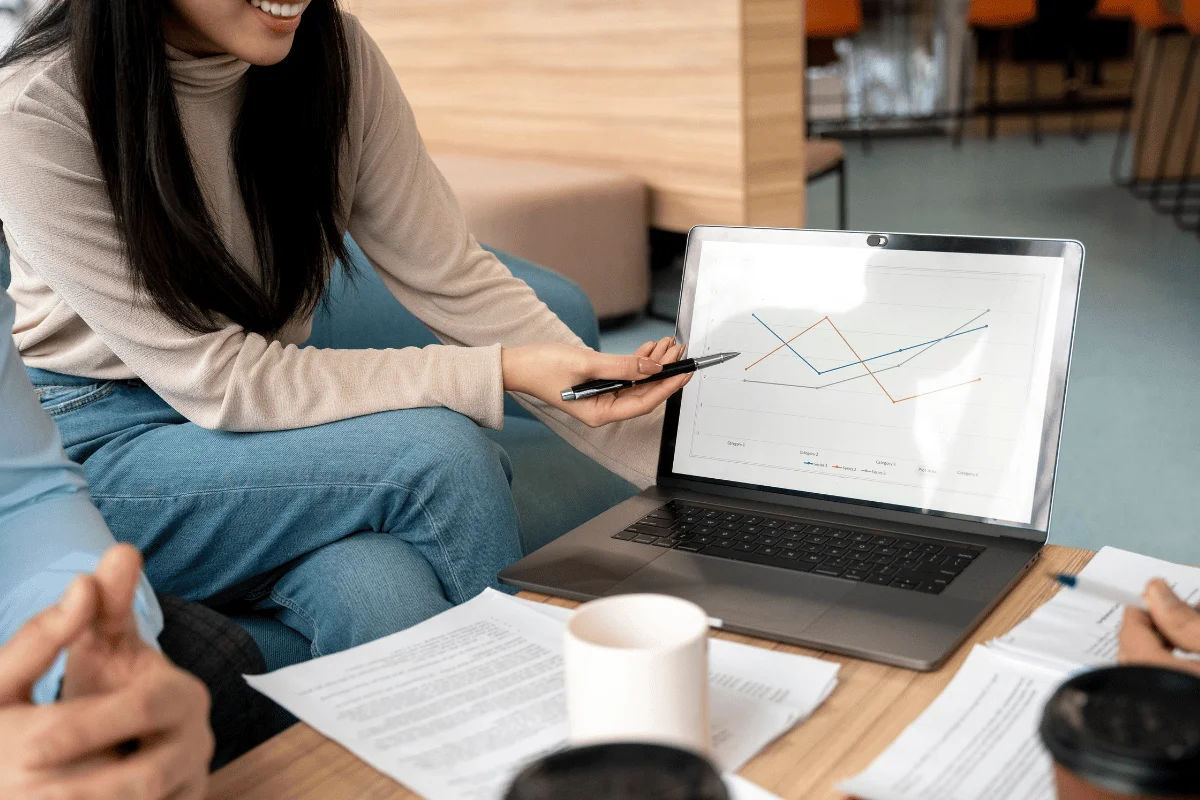 A woman points at a line graph on a laptop during a meeting, with papers and cups visible on the table.