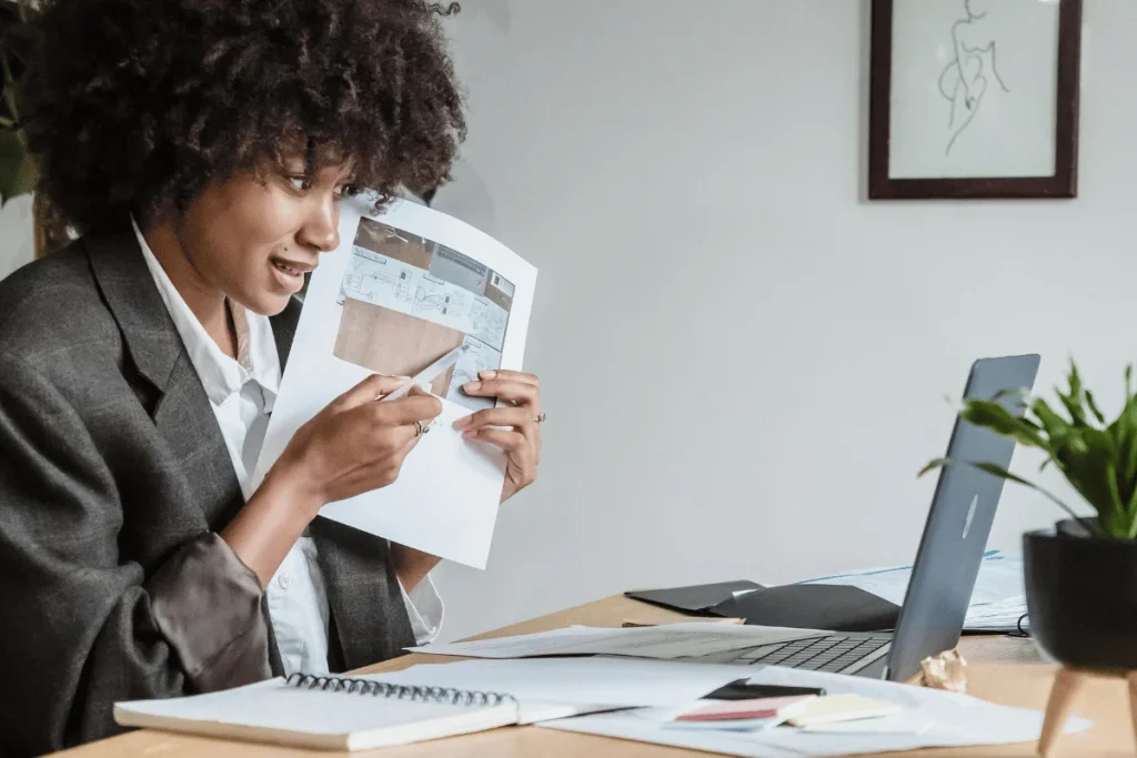 A person in a blazer examines blueprints at a desk cluttered with papers and a laptop, with a plant nearby.