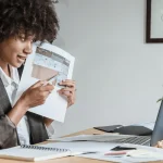 A person in a blazer examines blueprints at a desk cluttered with papers and a laptop, with a plant nearby.