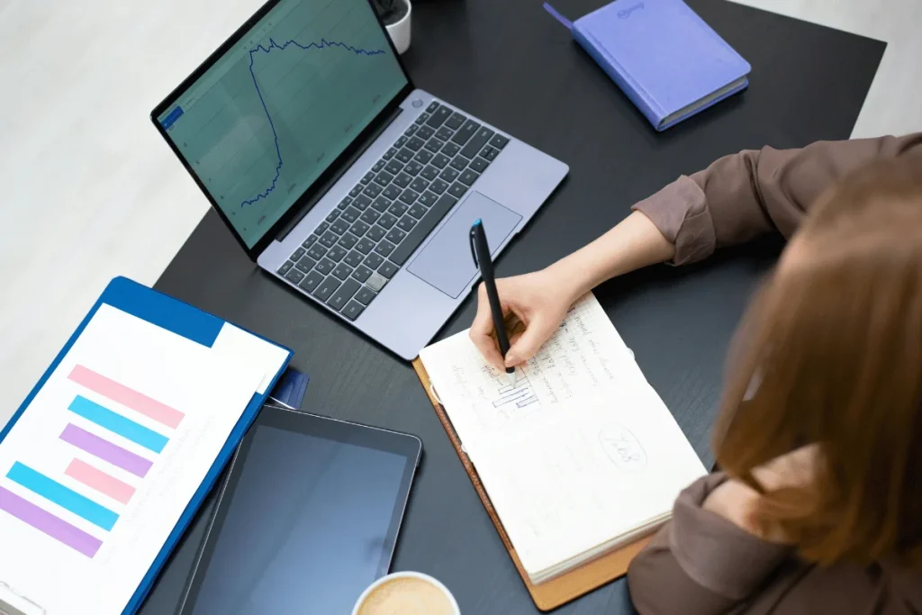 A person writes notes in a notebook while analyzing data on a laptop, surrounded by graphs, a tablet, and a coffee cup.