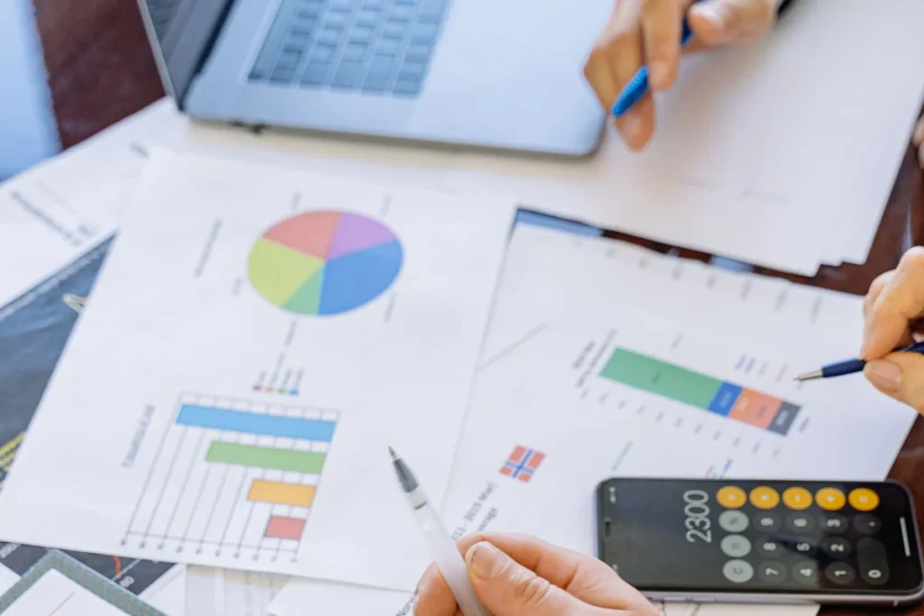 Hands holding pens are analyzing financial charts and graphs on papers spread across a table, with a laptop and a calculator displaying "2300."