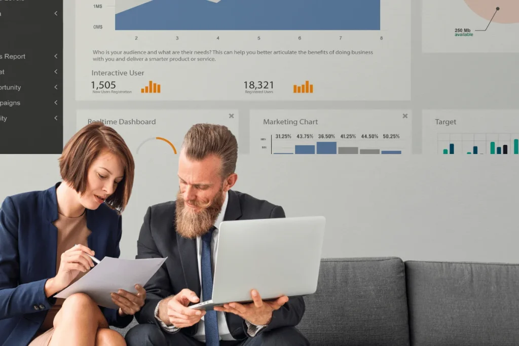 Two professionals analyze data on a laptop, surrounded by graphs and metrics displayed on a wall-mounted screen.