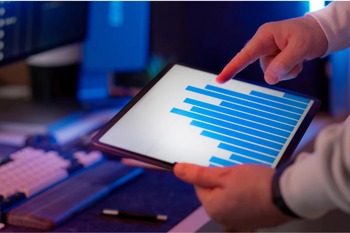 Hands hold a tablet displaying a blue bar graph in a dimly lit workspace, suggesting data analysis or technology focus.