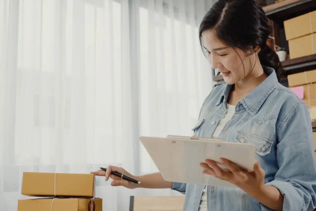 A person in a denim jacket examines a clipboard while standing next to several packed boxes in a bright room.