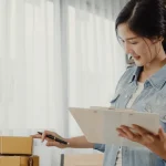 A person in a denim jacket examines a clipboard while standing next to several packed boxes in a bright room.