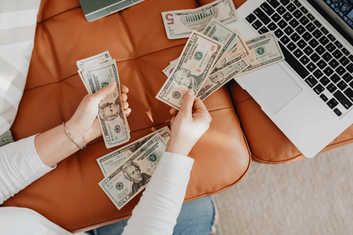 A person counting various U.S. banknotes on a leather couch, with a laptop visible in the background.