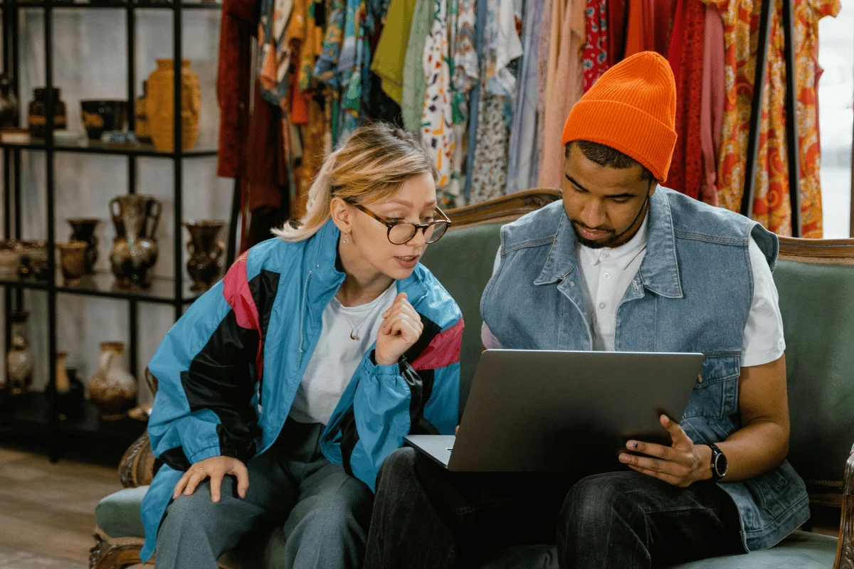 Two individuals sit together, focused on a laptop, with a colorful clothing rack and decorative items in the background.