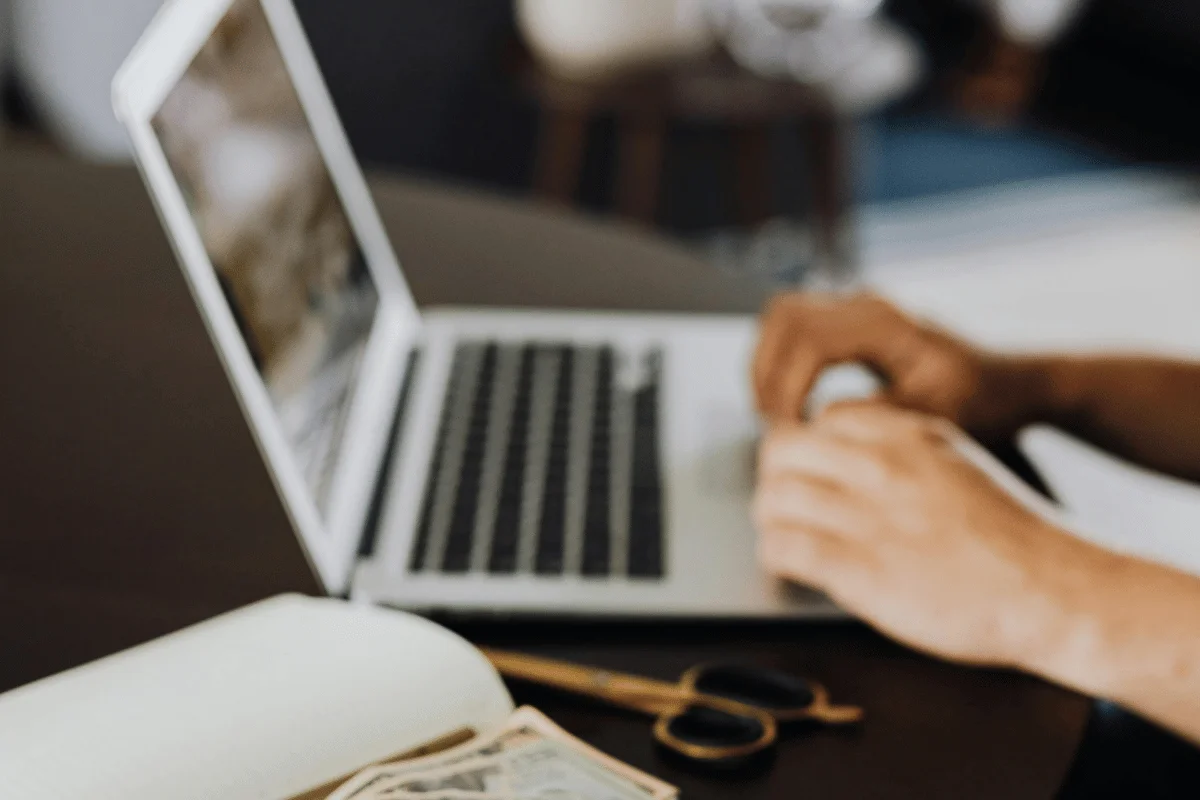 A person's hands typing on a laptop, with sunglasses and cash visible on a wooden table in a cozy workspace.