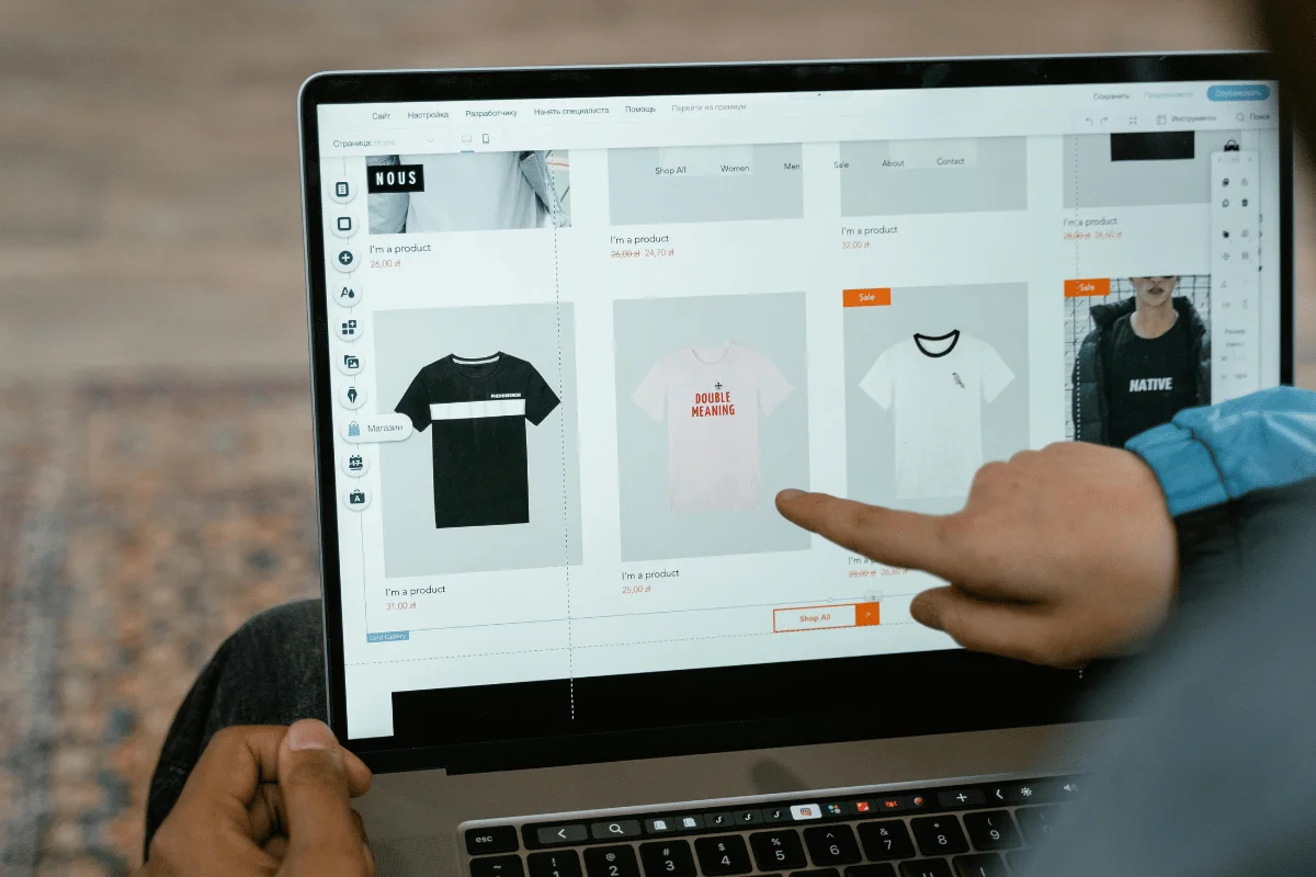 A person points at a laptop displaying a clothing website, showcasing various t-shirts for sale, including a pink one with "DOUBLE MEANING."