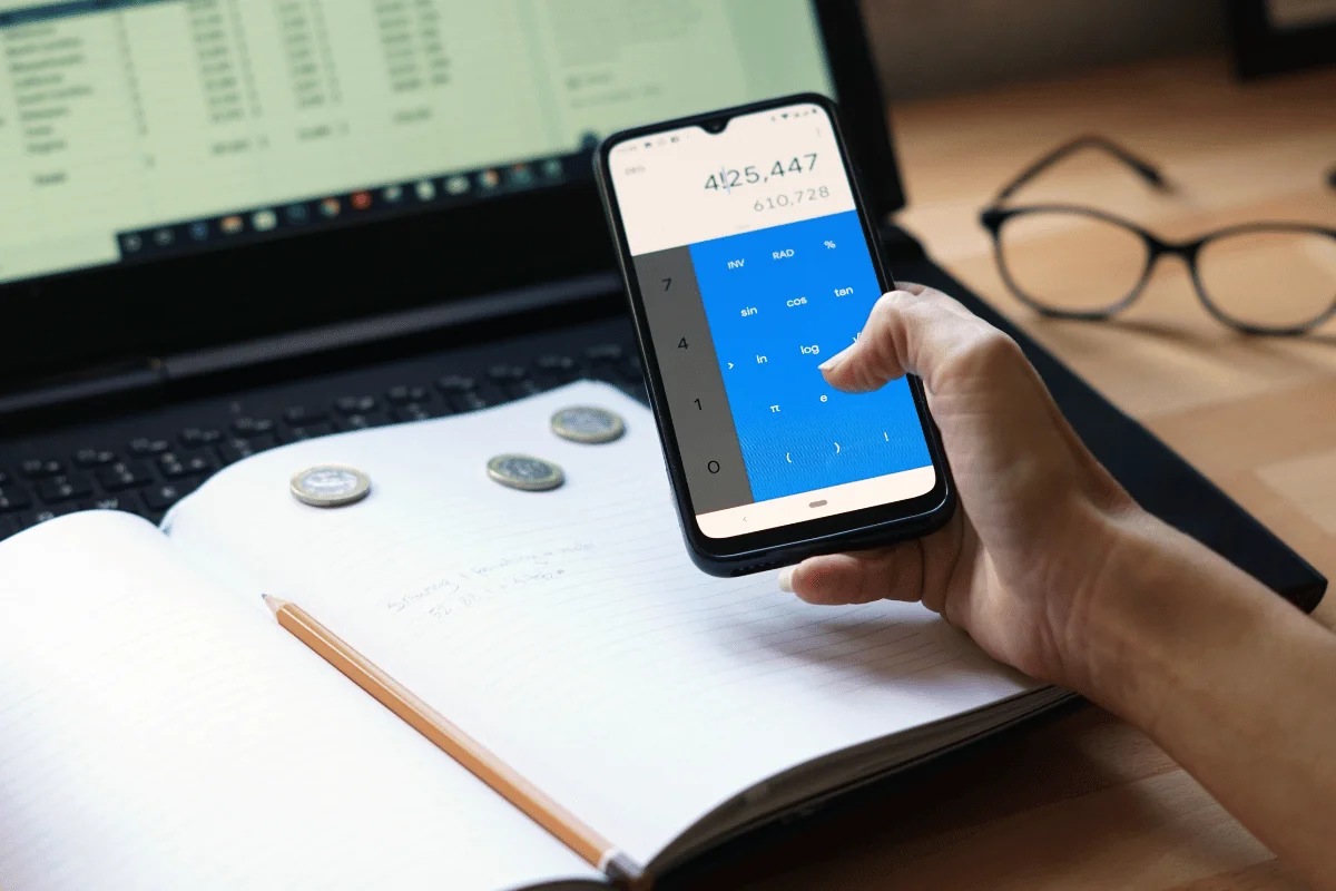 A hand holds a smartphone displaying a calculator app, beside a notebook, coins, and a laptop with a spreadsheet open.