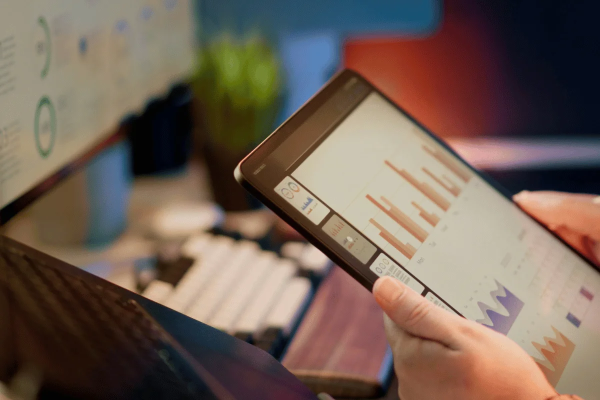 A person holds a tablet displaying colorful bar graphs and charts, with a computer monitor and keyboard in the background.