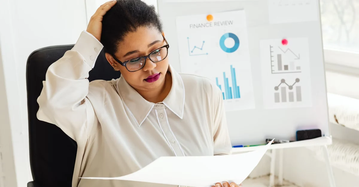 Woman reviewing financial documents at desk with marketing charts and graphs on whiteboard behind her.