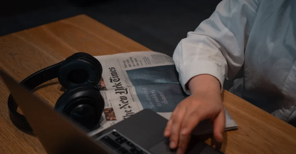 Person using ecommerce analytics tools on laptop while reading newspaper and wearing headphones at wooden desk.