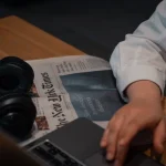Person using ecommerce analytics tools on laptop while reading newspaper and wearing headphones at wooden desk.
