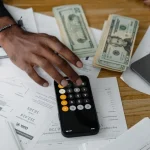 Ecommerce cash flow management workspace with calculator, dollar bills, financial documents, and laptop on wooden desk.