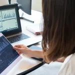 Woman analyzing ecommerce predictive analytics data on tablet and monitor at desk workspace.