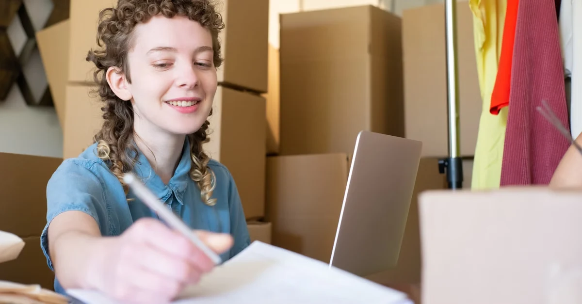 Young woman analyzing ecommerce marketing analytics on laptop while packing boxes for shipment.