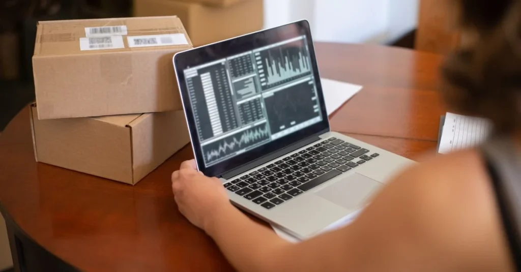 Person analyzing ecommerce marketing budget data on laptop next to cardboard shipping boxes on wooden desk.