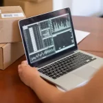 Person analyzing ecommerce marketing budget data on laptop next to cardboard shipping boxes on wooden desk.