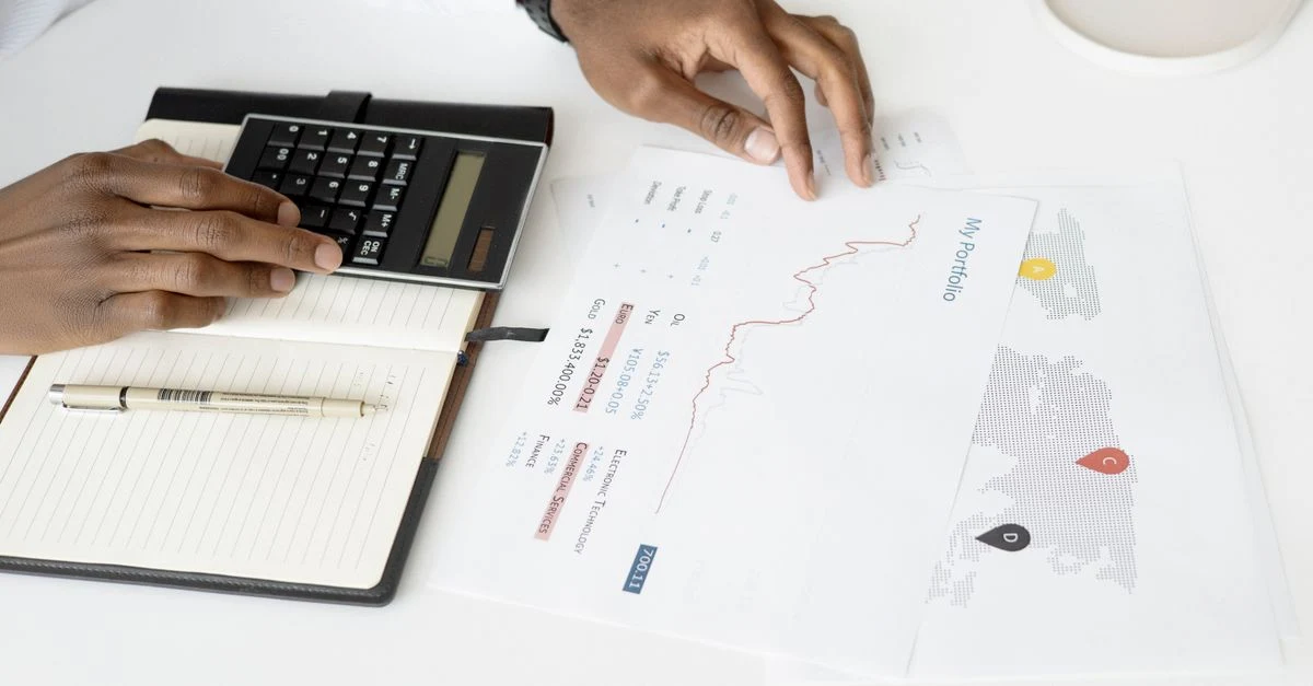 Person calculating ecommerce marketing budget using calculator and reviewing financial reports on white desk.